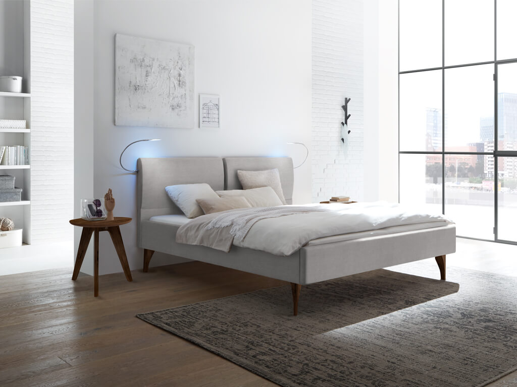 Ein graues Polsterbett mit geteiltem Kopfteil und dunklen Holzfüßen in einem hellen, modernen Schlafzimmer mit einem Teppich.