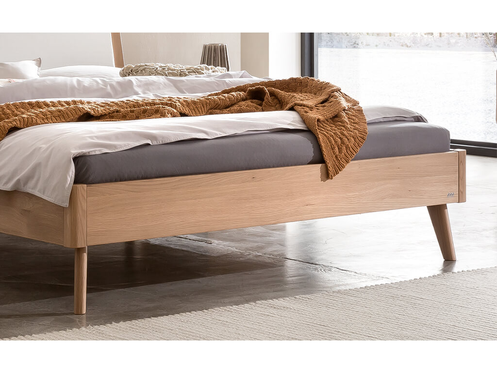 Eine Nahaufnahme des Fußendes eines Bettes mit einem hellen Eichenholzgestell. Man sieht die konisch zulaufenden Beine und ein kleines Logo auf der Seite des Bettrahmens. Das Bett steht auf einem grauen Boden mit einem hellen Teppich.