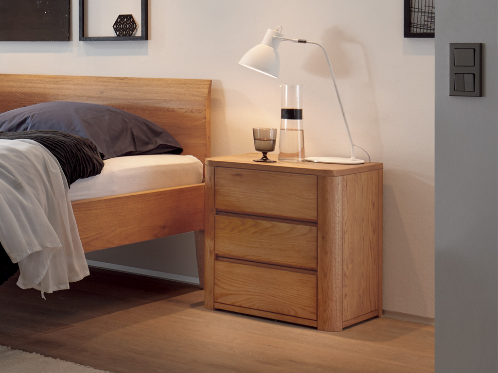 brauner Holznachttisch mit drei Schubladen und abgerundeten Seiten stehend neber einem Holzbett stilvoll dekoriert mit einer Nachttischlampe Koro von Hasena