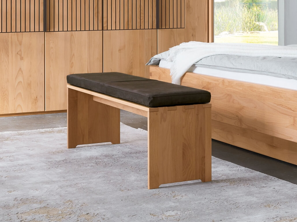 Wöstmann WSM 4400 - Sitzbank mit Bankkissen (1481) | Erle massiv | B: 120,0 cm Holzbank mit schwarzem Polster in modernem Schlafzimmer mit Holzboden und Bett