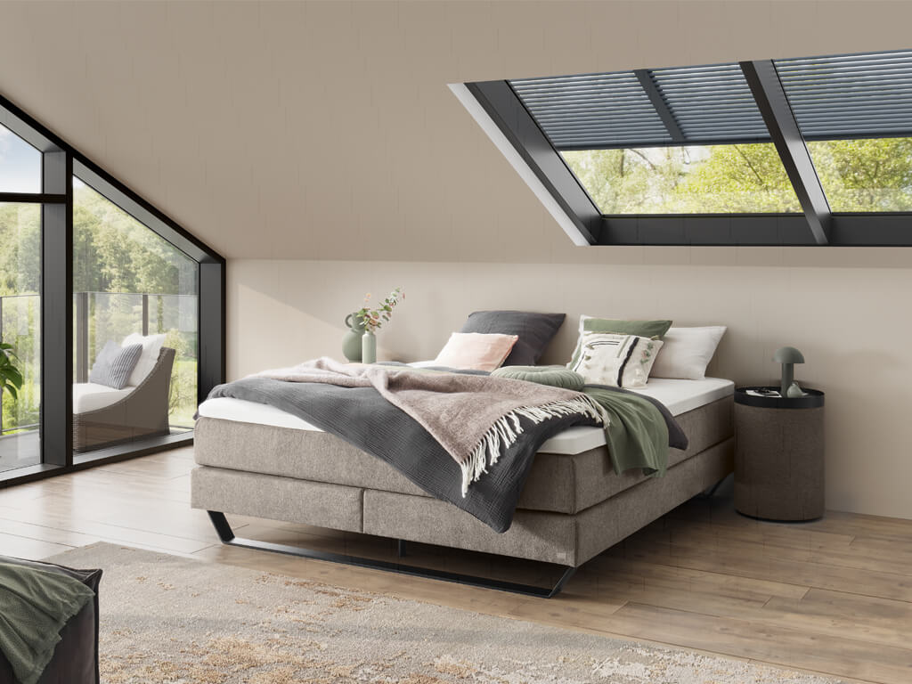 Ein Schlafzimmer, das ein Boxspringbett in einem Grauton mit verschiedenen Kissen und Decken zeigt, umgeben von großen Fenstern.