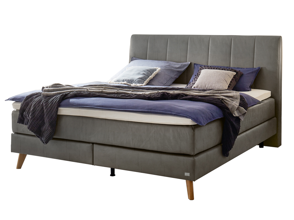 Ein Boxspringbett mit dunkelgrauem Kopfteil, Holzfüßen und passender Matratze und Bettwäsche vor einem weißen Hintergrund.