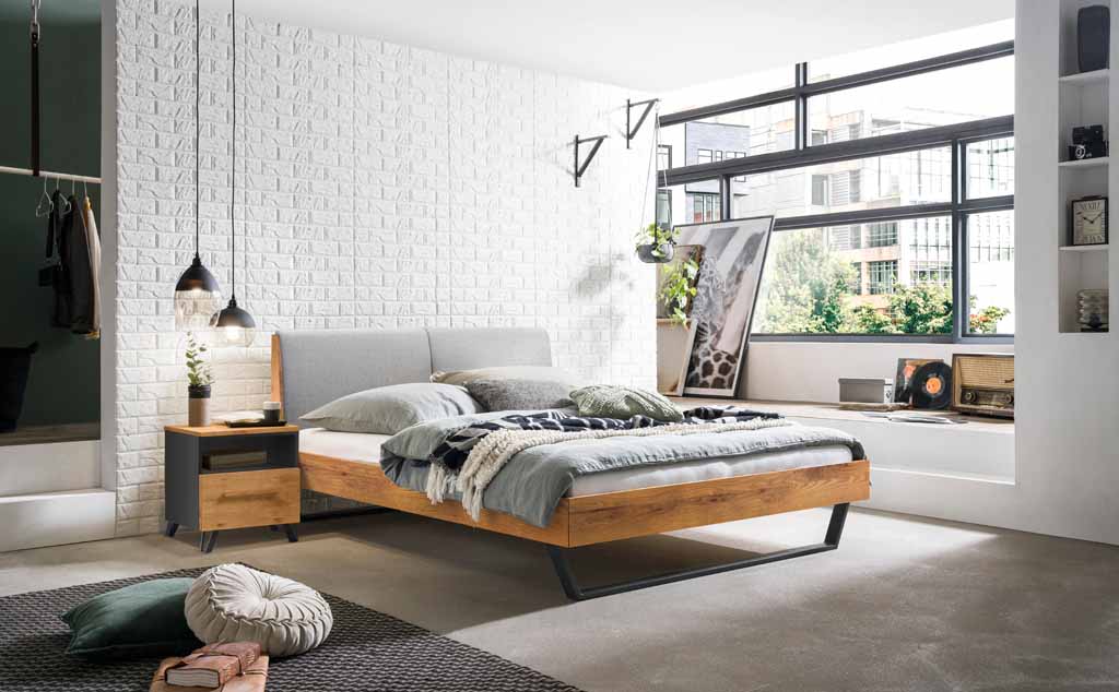 Hasena Trento - Bett Pietro | Wildeiche natur, geölt | Kopfteil Stoff grau Stilvolles Schlafzimmer mit einem Holzbett, weißen Ziegelwänden, großen Fenstern und dekorativen Elementen.