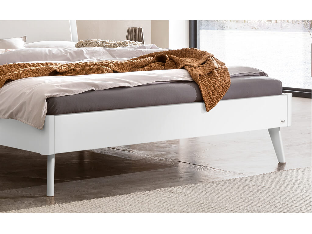 Eine Nahaufnahme des Fußendes eines Bettes mit einem weiß lackierten Bettgestell und passenden, konisch zulaufenden Beinen. Die Kanten des Rahmens sind abgerundet. Das Bett steht auf einem grauen Boden mit einem hellen Teppich.
