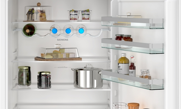 Innenansicht des Kühlabteils mit gut organisierten Lebensmitteln, darunter Kuchen, Desserts, Gurkengläser und Käse. Man sieht den gewellten Flaschenhalter und die seitlich verstellbaren Türablagen.