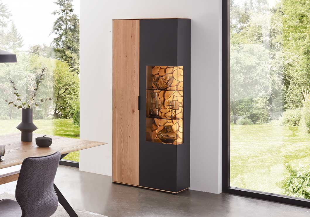 Wöstmann WM 2140 - Vitrine | Type 6843/6844 | B: 79,5 cm | Wildeiche massiv Ein moderner Schrank mit Glasvitrine, gefüllt mit dekorativen Gegenständen, steht in einem Raum mit großem Fenster und Blick auf einen Garten.