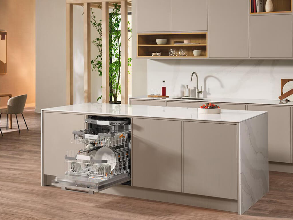 Eine moderne Küche mit einer geöffneten Miele Geschirrspülmaschine, die in die Kücheninsel integriert ist. Die Geschirrkörbe sind herausgezogen und mit Geschirr beladen.