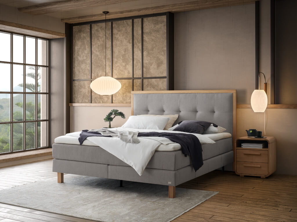 Ein Hasena Boxspringbett Pronto Solara Presley mit hellgrauem Stoffbezug in einem warm und stilvoll eingerichteten Schlafzimmer.