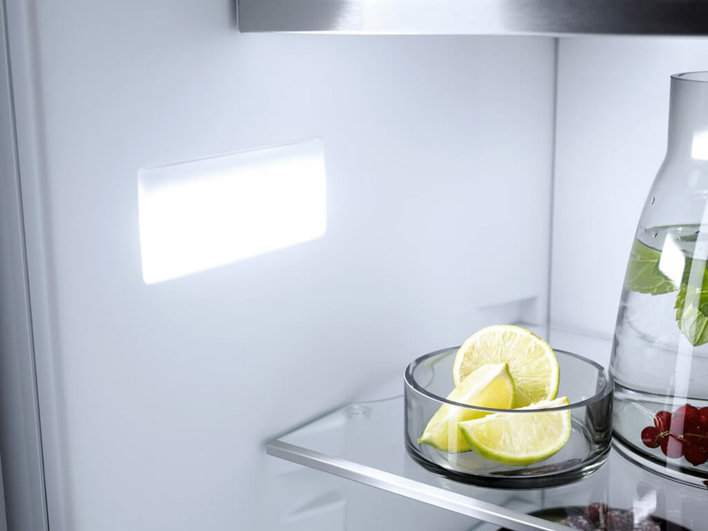 Eine Nahaufnahme des Innenraums eines Miele Kühlschranks. Man sieht ein beleuchtetes LED-Panel und eine Glasschale mit aufgeschnittenen Limetten auf einer Glasablage.