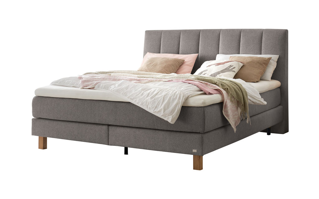 Ein Boxspringbett in einem hellen Grauton mit einem vertikal gesteppten Kopfteil, freigestellt auf weißem Hintergrund.