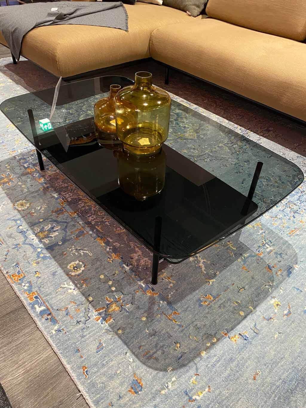 Ein eleganter Couchtisch mit Glasplatte, verziert mit amberfarbenen Vasen, steht auf einem gemusterten Teppich neben einem beigen Sofa.