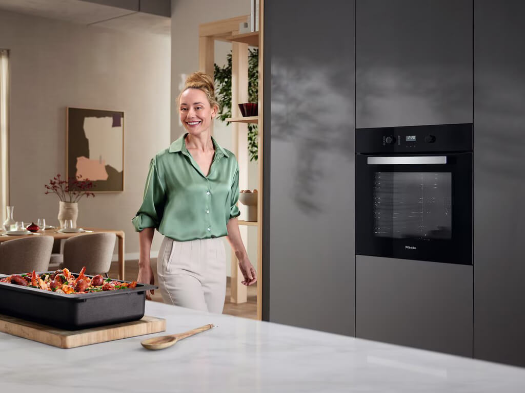 Eine lächelnde Frau in einer modernen Küche mit einem schwarzen Miele-Einbaubackofen. Im Vordergrund steht ein Backblech mit einem fertig gebratenen Huhn und Gemüse auf einer Arbeitsplatte.