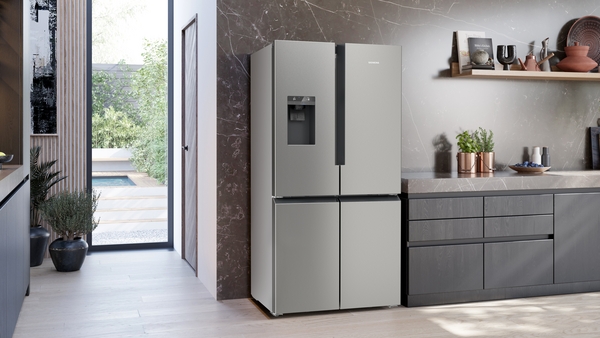 Ein eleganter, silberner French-Door-Kühlschrank steht in einer modernen Küche mit grauen Schränken und einem Fenster mit Blick auf einen Garten.