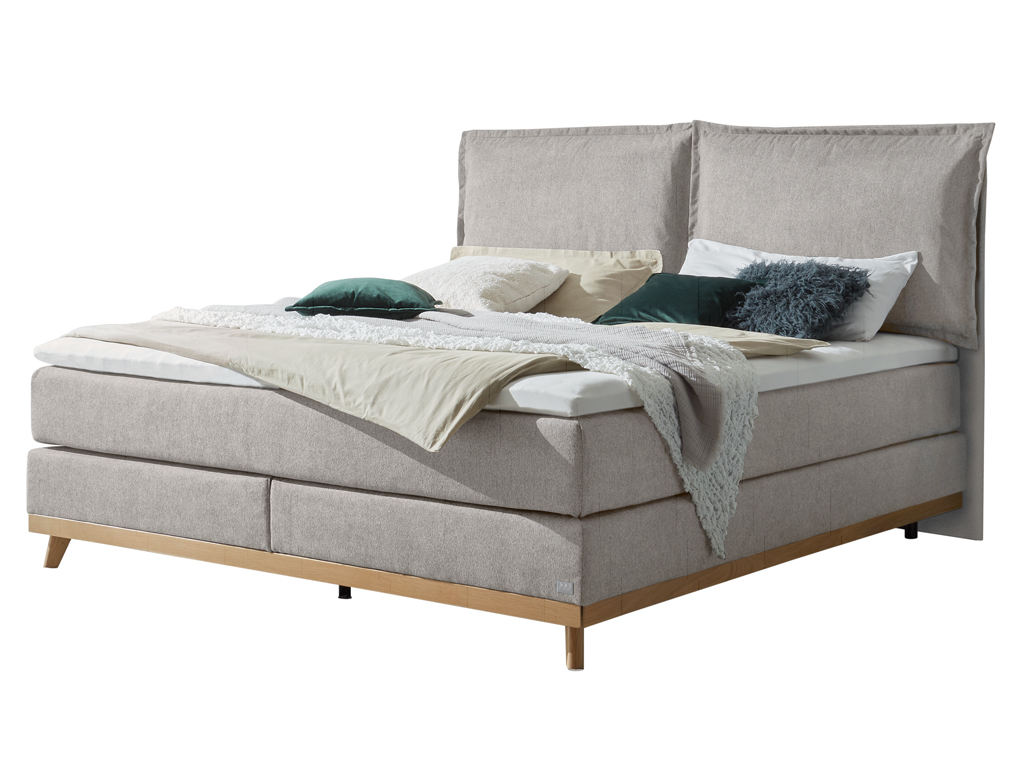 Ein elegantes Boxspringbett in einem helleren Grauton mit einem gepolsterten Kopfteil und Holzfüßen, geschmackvoll dekoriert mit Kissen und einer Decke auf weißem Hintergrund.