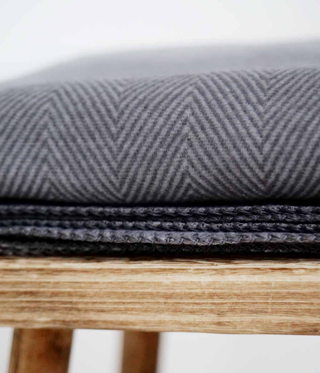 RIEMA. FINN – Wohndecke, Bio-Baumwolle - Fischgrät | 140 x 200 cm | dunkelgrau Nahaufnahme eines grauen Herringbone-Stuhlpolsters auf einem Holzhocker.