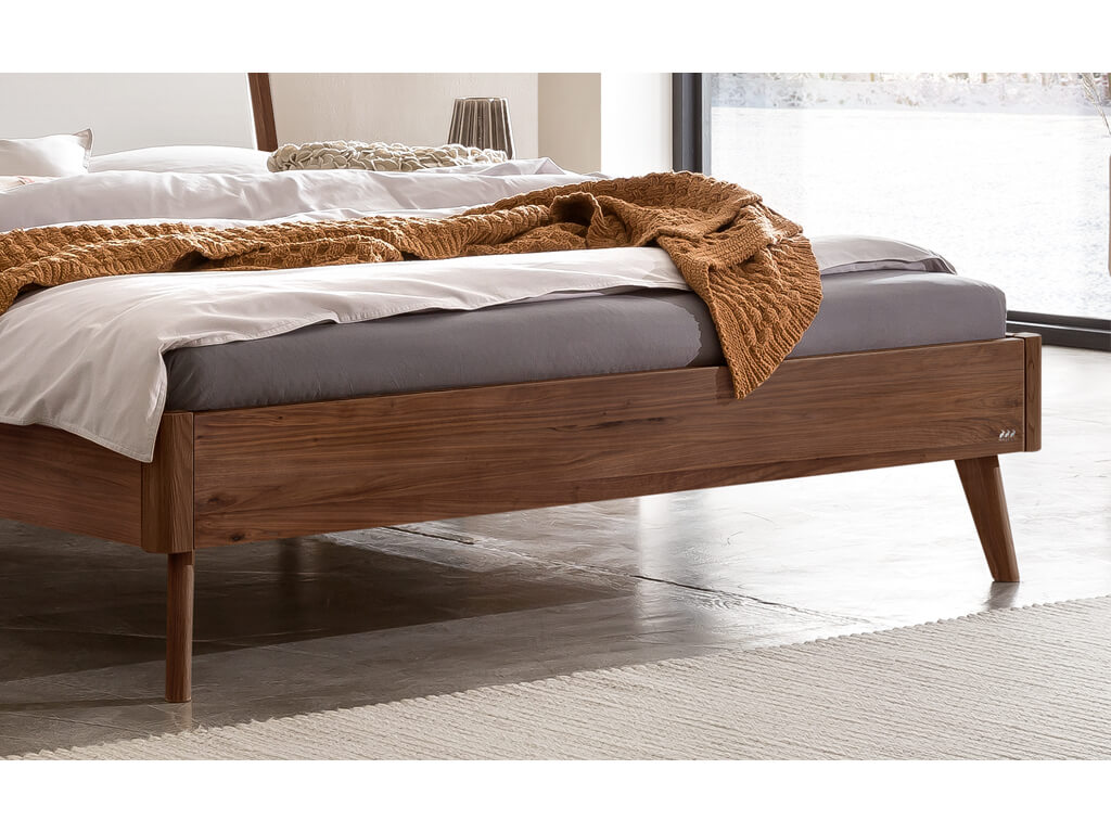 Eine Nahaufnahme des Fußendes eines Bettes aus dunklem Walnussholz. Man sieht die konisch zulaufenden Beine und ein kleines Logo auf der Seite des Bettrahmens. Das Bett steht auf einem grauen Boden mit einem hellen Teppich.