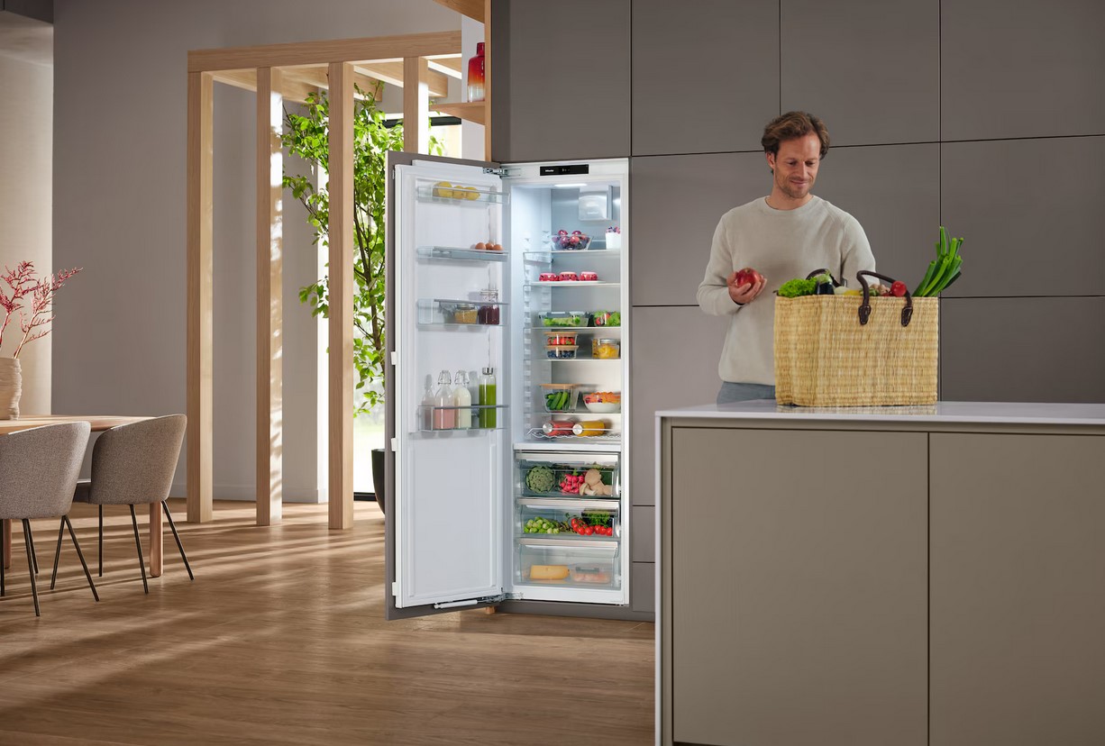 Ein lächelnder Mann steht in einer hellen Küche neben einem offenen Miele Kühlschrank, der mit frischen Lebensmitteln gefüllt ist. Er hält einen Apfel in der Hand und neben ihm steht ein geflochtener Korb mit Gemüse.
