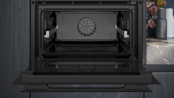 Frontale Detailansicht des geöffneten Siemens Backofen-Innenraums. Fokus auf die schwarze Emaillierung, das zentrale 4D-Heißluft-Gebläse und die seitlichen Aufnahmegitter für Backbleche bei vollständig heruntergeklappter Glastür.