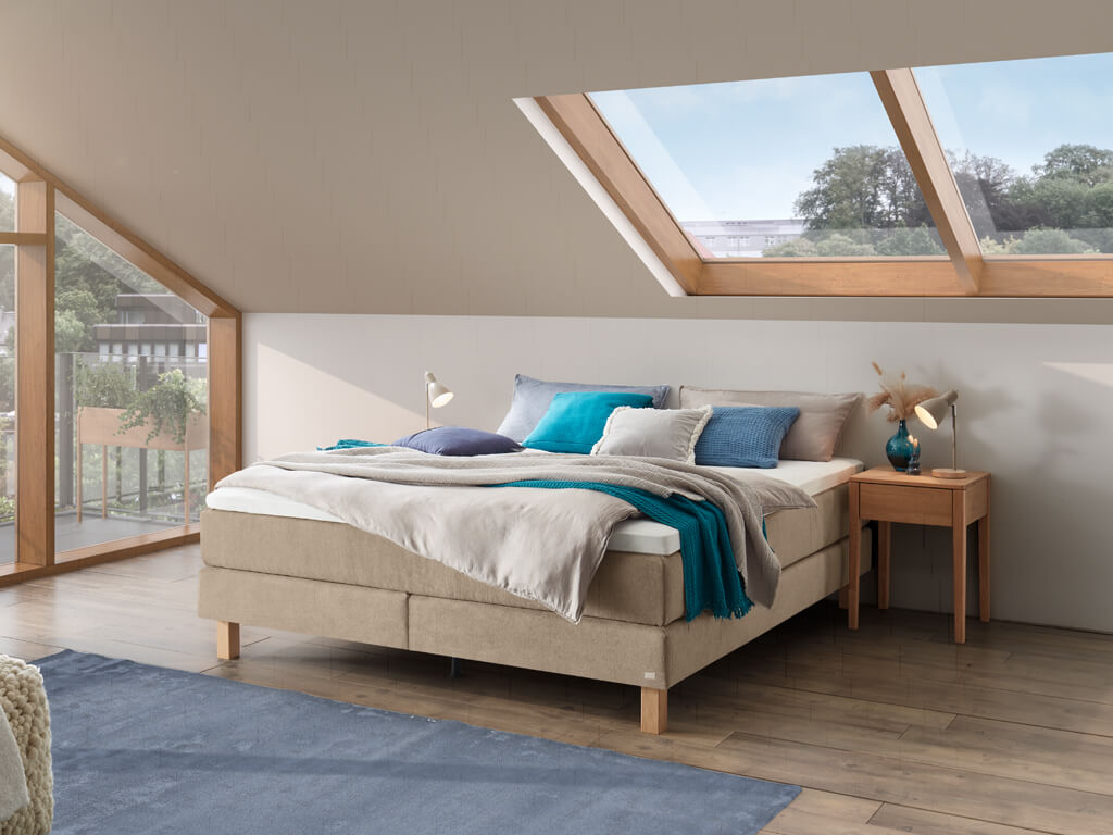 Ein helles Schlafzimmer in einem Dachgeschoss mit dem sandfarbenen Hasena Boxspringbett "Pronto Carlota", das unter einem großen Dachfenster steht.