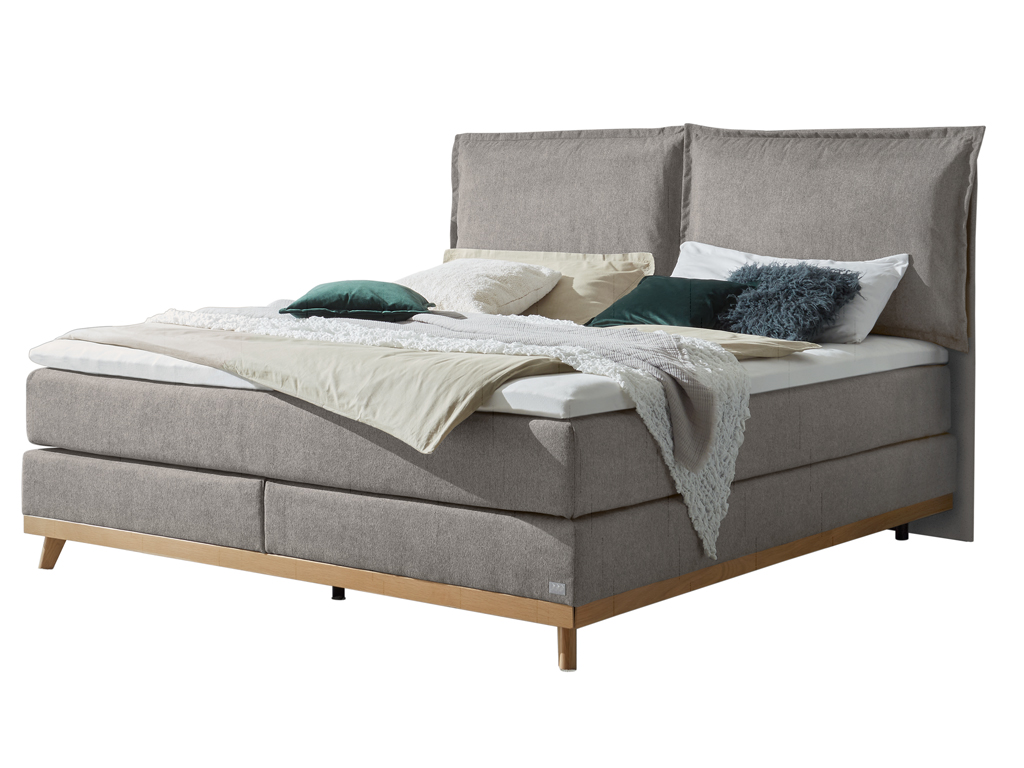Ein stilvolles Boxspringbett in einem Grauton mit einem gepolsterten Kopfteil und Holzfüßen, dekoriert mit Kissen und einer Decke auf weißem Hintergrund.