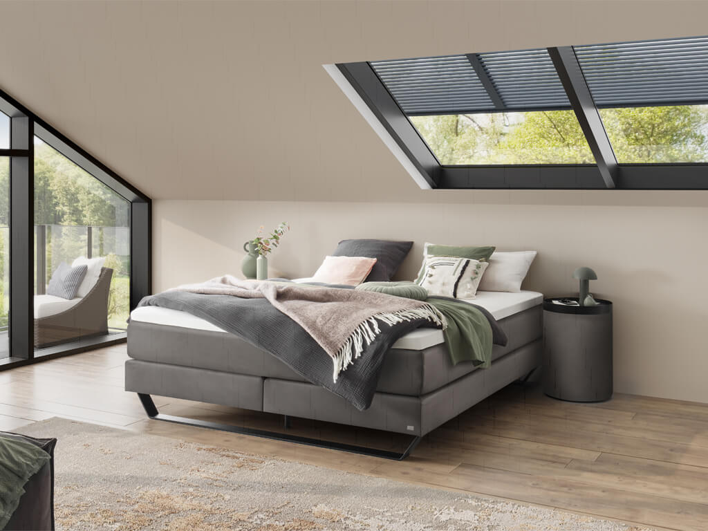 Ein modern eingerichtetes Schlafzimmer mit einem grauen Boxspringbett und einem großen Fenster, das eine natürliche Umgebung überblickt.