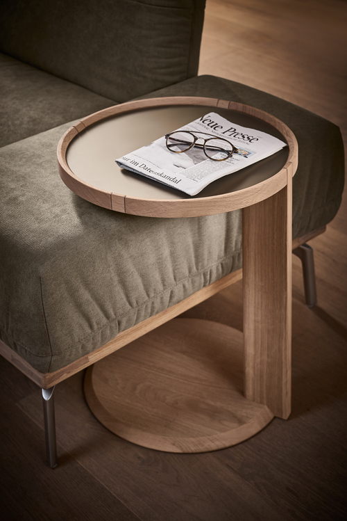 Wimmer Signatura | Beistelltisch mit Glaseinsatz | 309100 Ein moderner Beistelltisch aus hellem Holz neben einem Sofa, darauf liegen eine Zeitung und eine Brille.