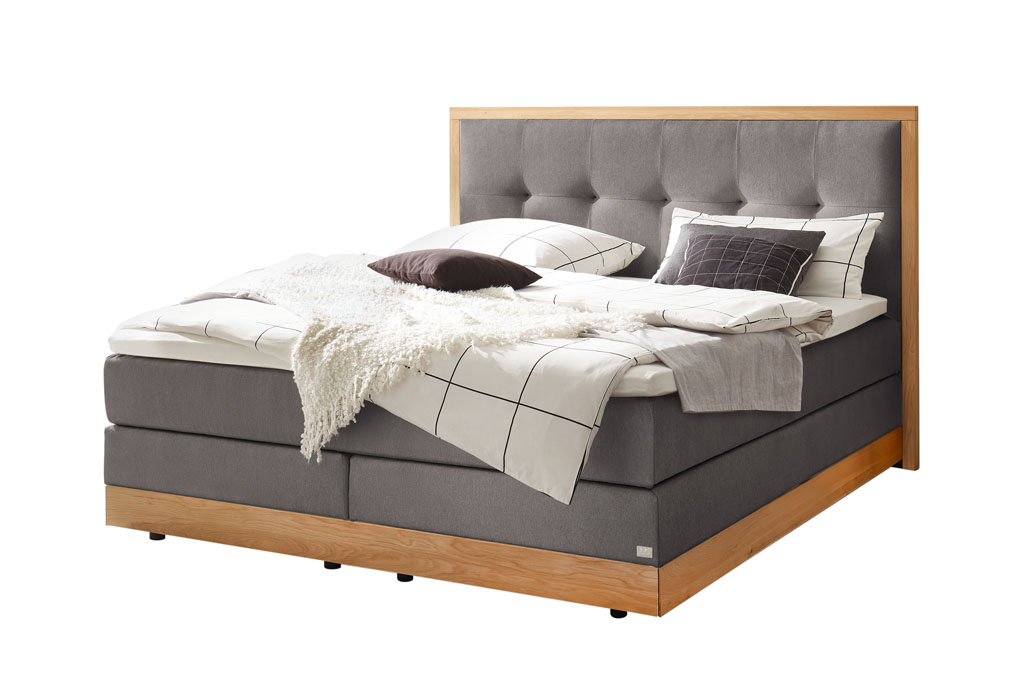 graues Boxspringbett mit braunem Holzrahmen, stilvoll dekoriert mit Decken und Kissen vor weißem Hintergrund