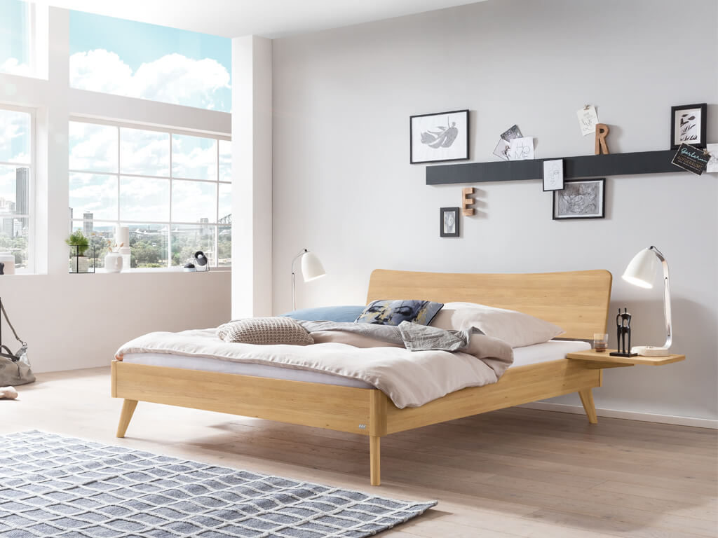 Ein Hasena Bett Caluso aus hellem Eichenholz ("Eiche bright") in einem hellen, modern eingerichteten Schlafzimmer mit großen Fenstern. Das Bett hat ein geschwungenes Kopfteil und schräg ausgestellte Beine.