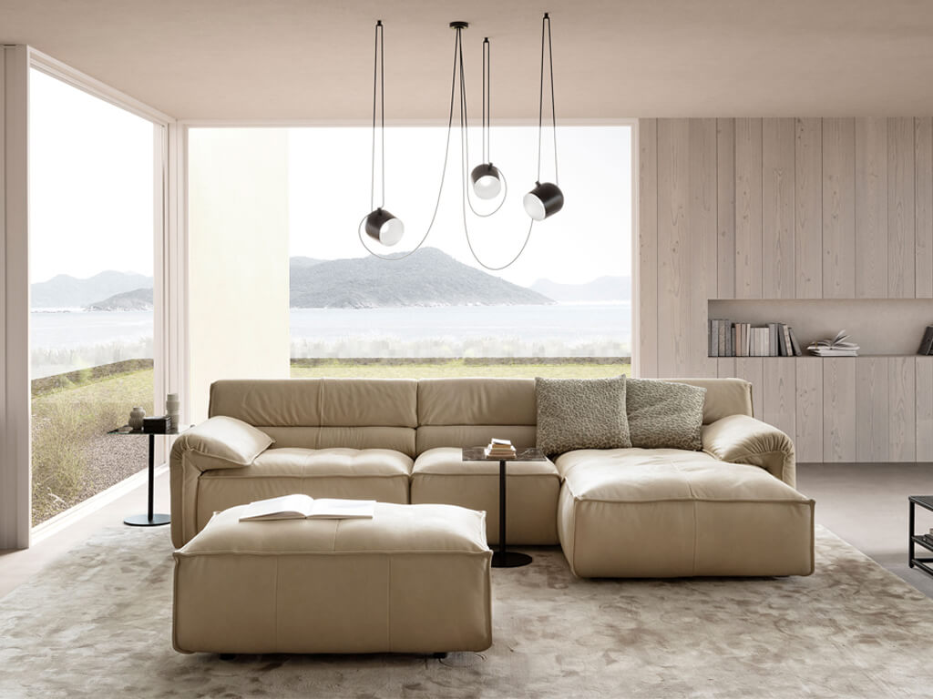 Ein modernes Wohnzimmer mit einem cremefarbenen Ecksofa und einem passenden Hocker, dekoriert mit grünen Zierkissen. Große Fenster bieten einen Blick auf eine Küstenlandschaft mit Bergen und Wasser.