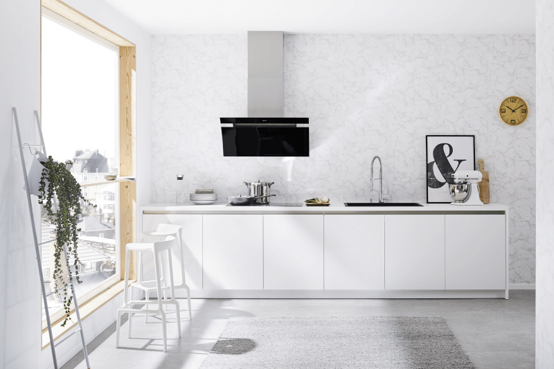 Eine minimalistische, helle Küche mit weißen Schränken, einem großen Fenster und einem hellen Teppich. Eine schwarz-silberne Dunstabzugshaube ist über einem schwarzen Kochfeld und Spülbecken angebracht. Zwei weiße Barhocker sind links zu sehen.