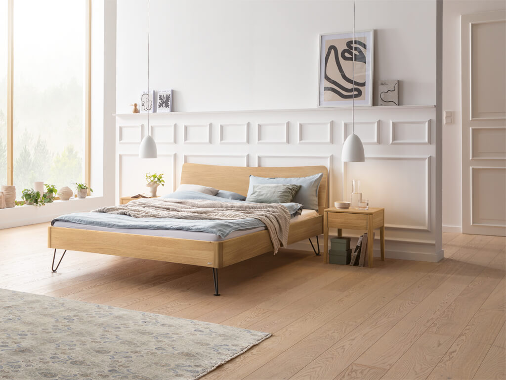 Ein Hasena Bett Locarno in heller Eiche (Eiche bright), in einem hellen Schlafzimmer mit minimalistischer Dekoration. Die schwarzen, anthrazitfarbenen Haarnadel-Beine sind sichtbar. Auf der linken und rechten Seite stehen passende Nachttische.