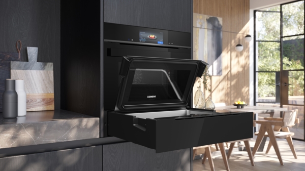 Eingebauter Siemens StudioLine Backofen mit Touch-Display über einer offenen, ausziehbaren schwarzen Schublade, die wie ein Dampfgarer aussieht. Die Klappe ist nach oben geöffnet. Die Szene ist eine moderne Küche mit viel Tageslicht.