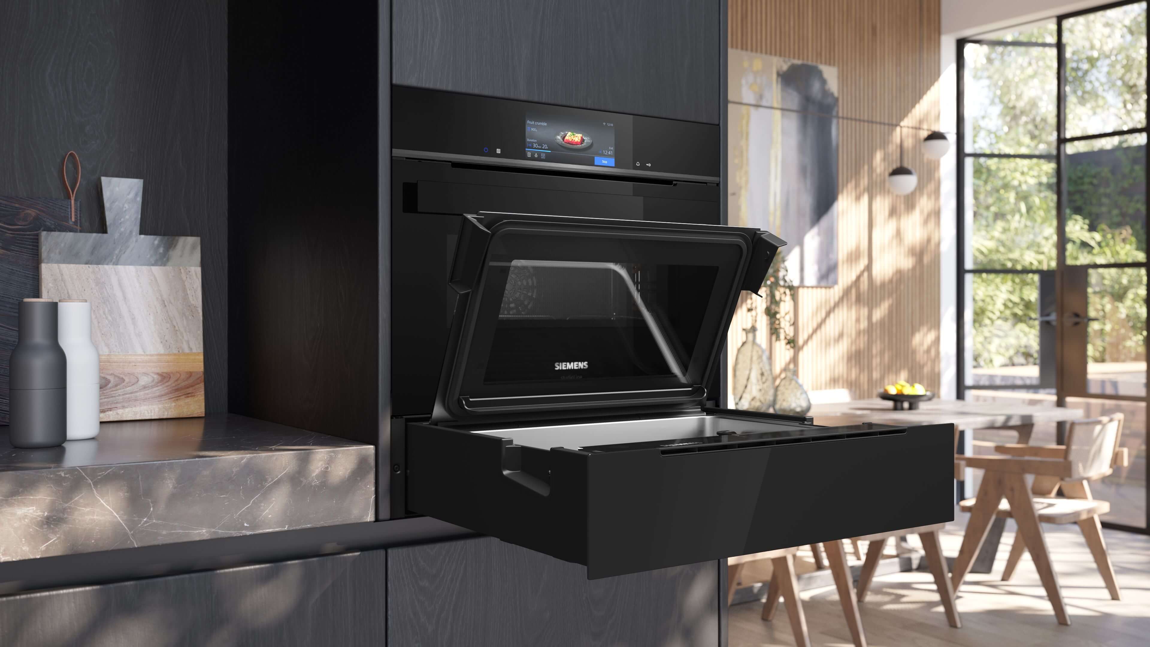 Eingebauter Siemens StudioLine Backofen mit Touch-Display über einer offenen, ausziehbaren schwarzen Schublade, die wie ein Dampfgarer aussieht. Die Klappe ist nach oben geöffnet. Die Szene ist eine moderne Küche mit viel Tageslicht.