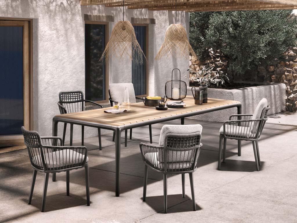 Rolf Benz Yoko 250 - Esstisch 220 x 100 cm Ein stilvoller Gartentisch aus Holz und Metall, umgeben von sechs passenden Stühlen, steht auf einer sonnigen Terrasse vor einem weißen Gebäude.