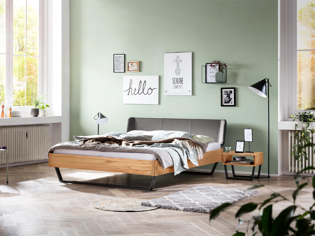 Ein Schlafzimmer mit einem Holzbett aus hellem Wildbuchenholz. Der Raum hat mintgrüne Wände und das Bett ist mit einem grauen, gepolsterten Kopfteil und einem schwarzen Metallgestell ausgestattet. Zwei kleine Teppiche liegen auf dem Holzboden.