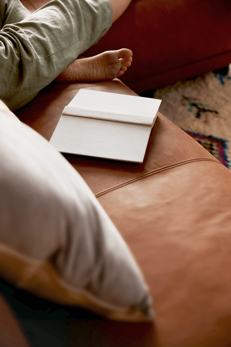 Ein geöffnetes Buch liegt auf einem Ledersofa, daneben eine entspannte Person mit Kreuzbeinen.