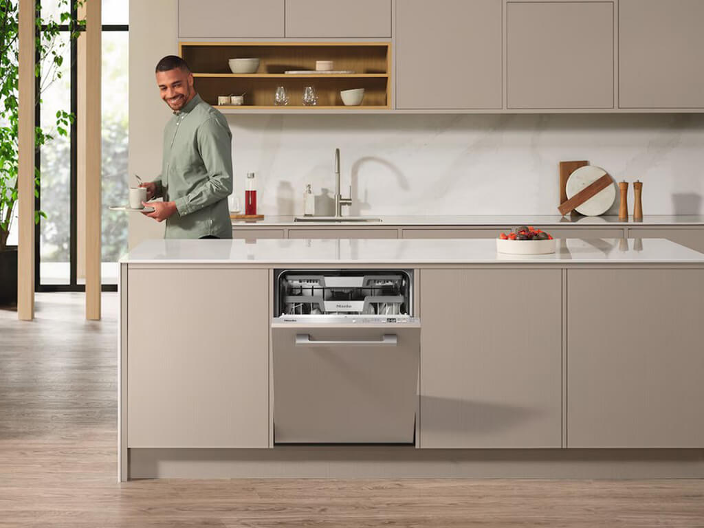 Ein lächelnder Mann steht in einer modernen Küche mit einer Geschirrspülmaschine in der Kücheninsel, die leicht geöffnet ist. Die Küche hat helle Schränke, eine weiße Arbeitsplatte und Holzboden.