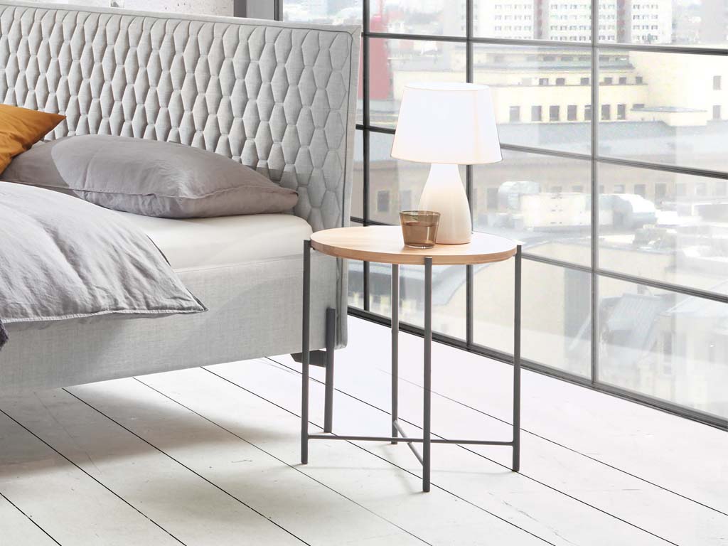 Kleiner brauner beistelltisch mit runder platte auf einem vierfüßigen metallgestell in grau stilvoll dekoriert neber einem bett in einem hellen zimmer nali von hasena