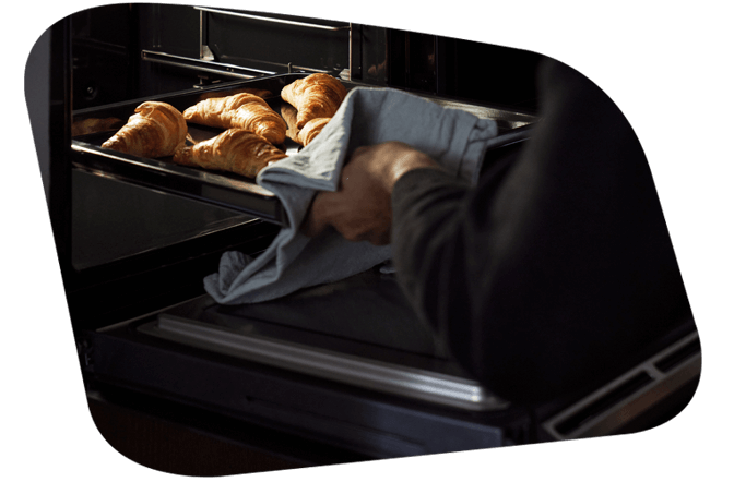 Eine Person entnimmt mit einem Küchentuch ein Blech mit frischen Croissants aus einem Siemens Dampfbackofen.