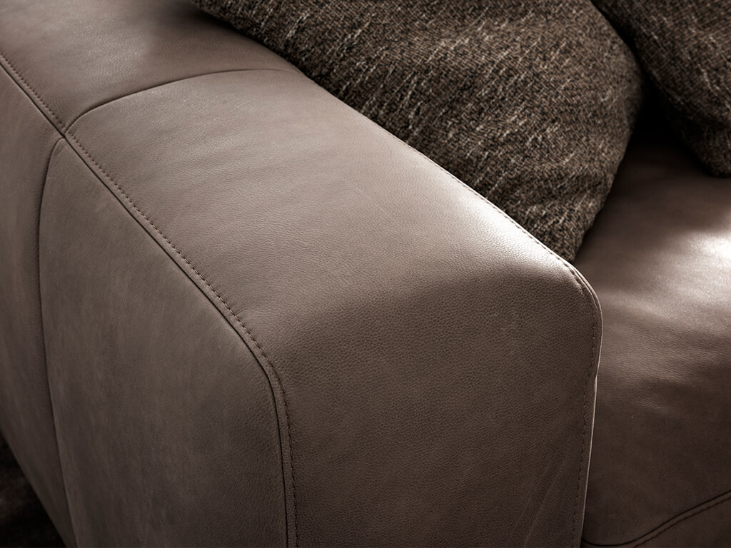 Nahaufnahme der Armlehne eines braunen Ledersofas mit sichtbaren Ziernähten und einem strukturierten Kissen im Hintergrund.
