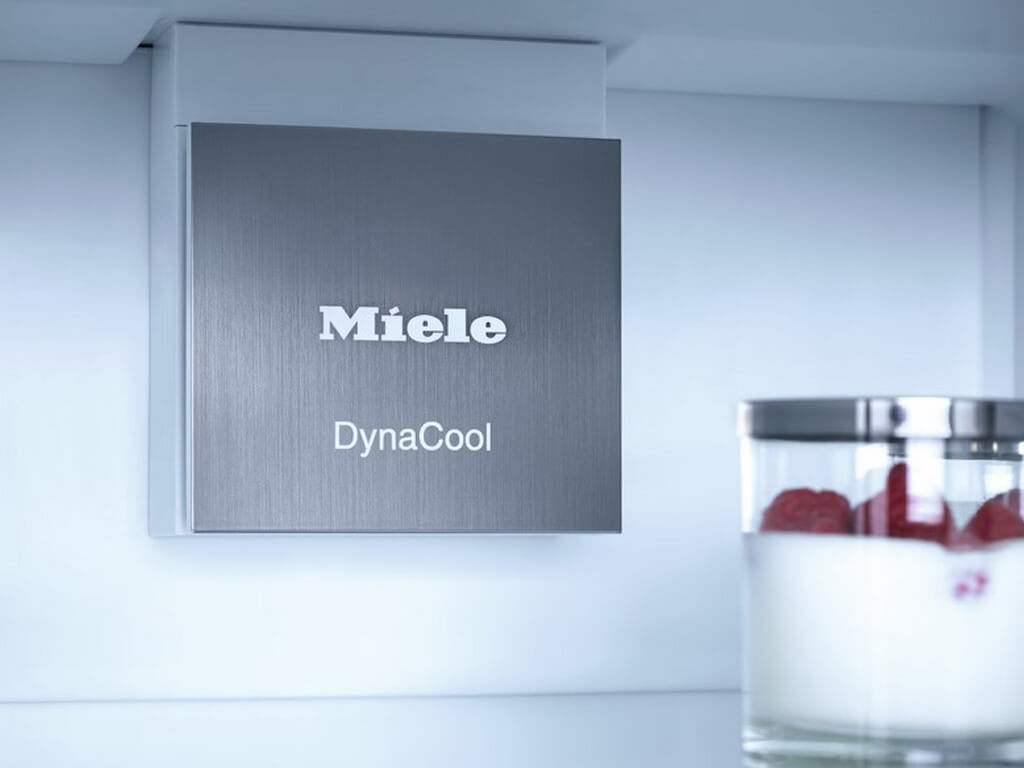 Das Bild zeigt das Innere eines Miele Kühlschranks. Man sieht eine silberne Blende mit den Logos "Miele" und "DynaCool", die sich oben an der Rückwand des Kühlschranks befindet.