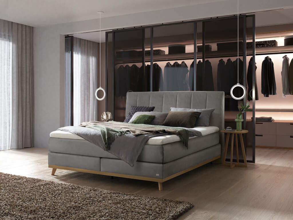 Ein Hasena Boxspringbett Pronto Stefania Roy mit einem stahlgrauen Stoffbezug, das sich harmonisch in ein modernes Schlafzimmer mit einem offenen Schrankbereich einfügt.