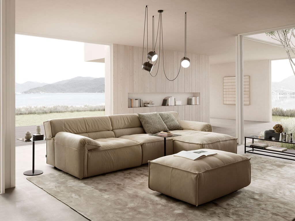 Ein modernes Wohnzimmer mit einem cremefarbenen Ecksofa und einem passenden Hocker, dekoriert mit grünen Zierkissen. Große Fenster bieten einen Blick auf eine Küstenlandschaft mit Bergen und Wasser, ähnlich wie in M1, aber aus einem leicht anderen Winkel.