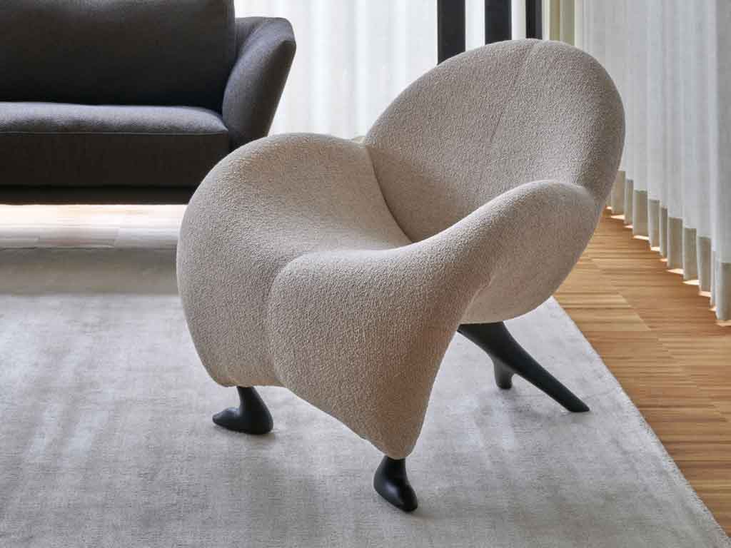Leolux Sessel | Papageno 931 Ein eleganter, moderner Designer-Sessel mit geschwungenem Design und weißem Stoffbezug, platziert auf einem Teppich neben einem Sofa in einem hellen Raum.