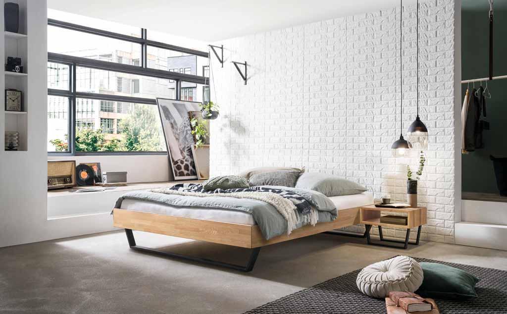 Hasena Trento - Bett Ostino | Eiche bianco, geölt Ein helles, modernes Schlafzimmer mit einem Holzplattformbett, dekoriert mit Pflanzen und skandinavischen Elementen, das auf einem Betonboden steht. Große Fenster lassen natürliches Licht herein.