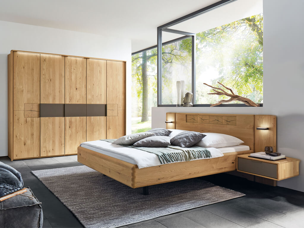 Wöstmann WSM 1600 - Schlafzimmer (Vorzugskombination) | Wildeiche massiv | Schrank 5-türig, Bett 180 x 200 cm, 2 Konsolen Ein modernes Schlafzimmer mit einem Bett und Kleiderschrank aus Holz, großen Fenstern und stilvollen Dekorationen.