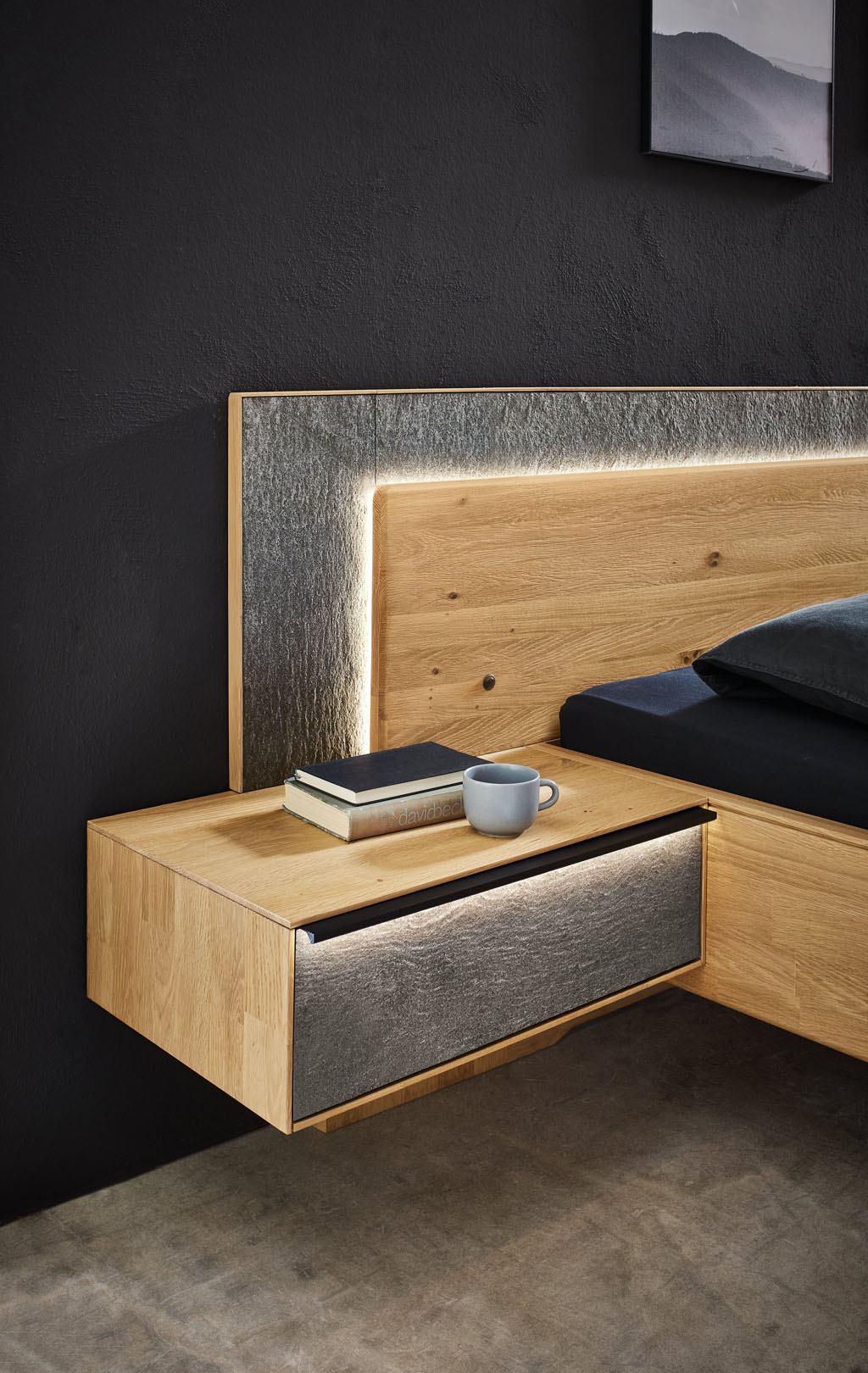 Detailaufnahme eines Bettes mit beleuchtetem Holzkopfteil und schwarzer Metallablage einer beleuchteten Hängekonsole mit grauer Front sowie einer grauer Kopfteilumrandung stilvoll dekoriert