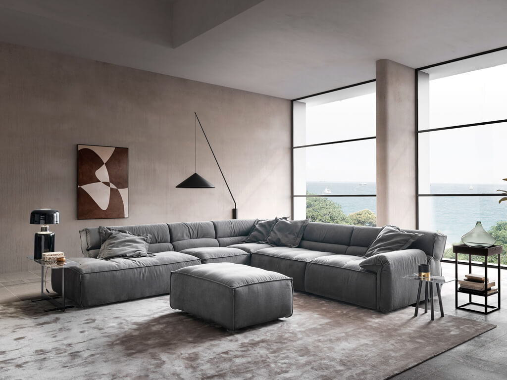 Ein modernes Wohnzimmer mit einem cremefarbenen Ecksofa und einem passenden Hocker, dekoriert mit grünen Zierkissen. Große Fenster bieten einen Blick auf eine Küstenlandschaft mit Bergen und Wasser.
