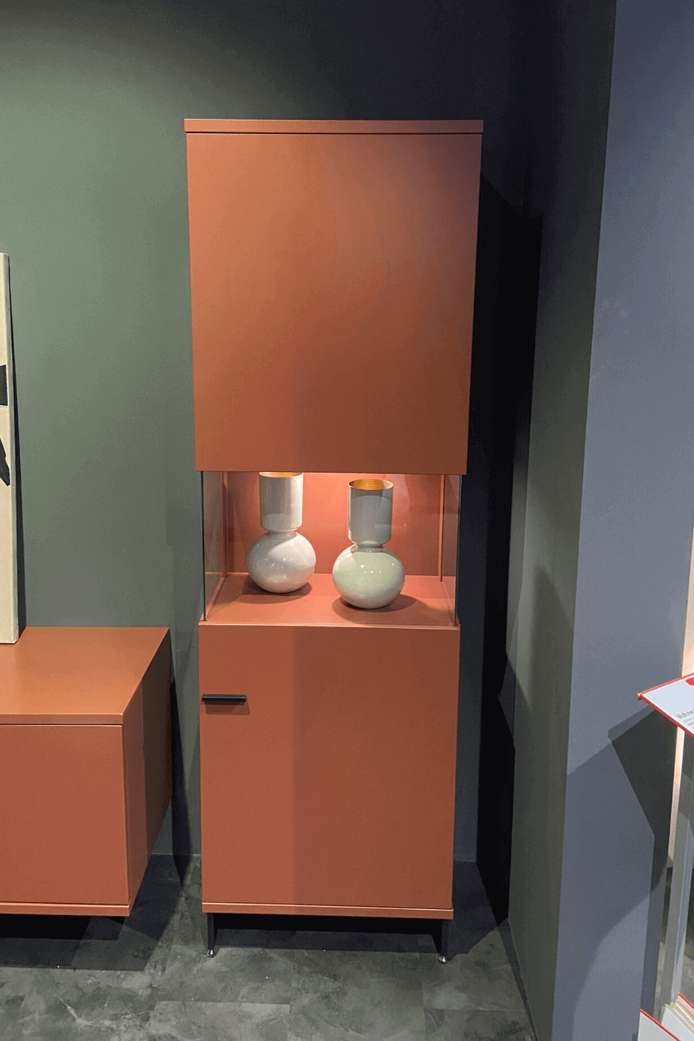 RMW LAVITA | Vitrine incl. Fußschalter Ein moderner Schrank in Terrakottafarbe mit einer integrierten Glasvitrine, darin stehen zwei weiße Vasen, im Hintergrund eine graue Wand.