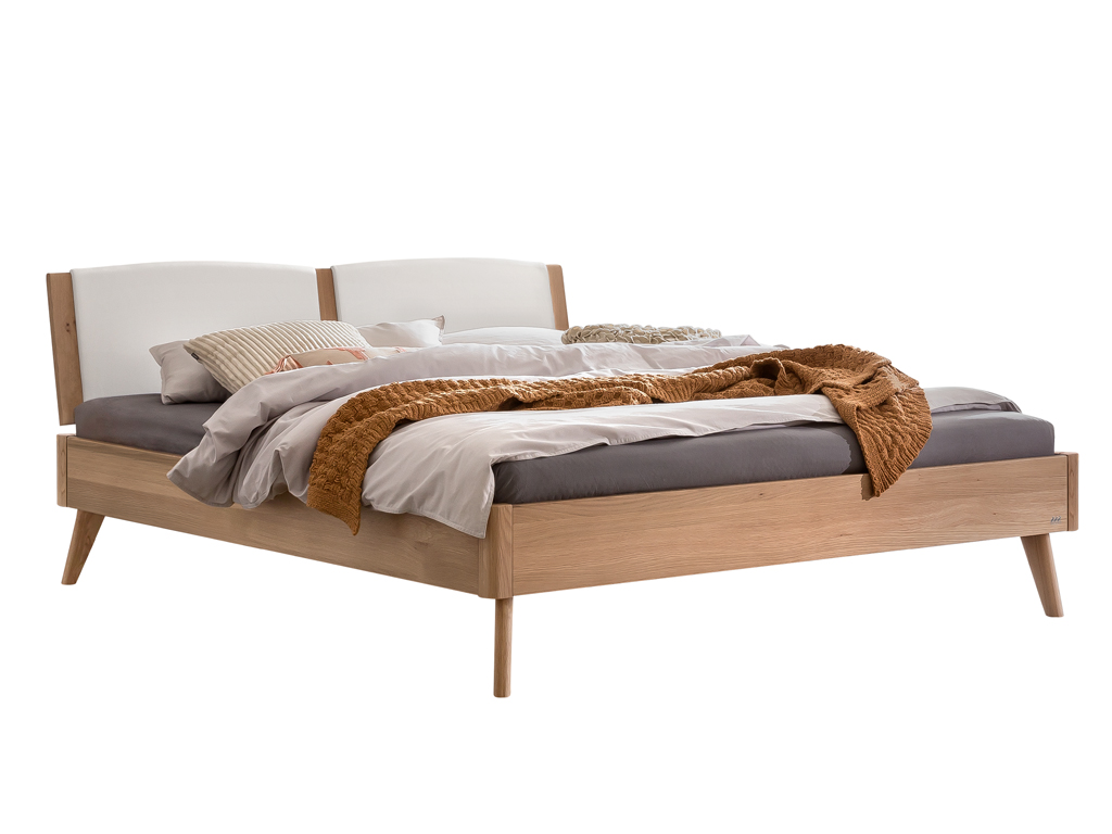 Ein stilvolles, modernes Doppelbett aus hellem Eichenholz mit einem gepolsterten, geteilten Kopfteil in Weiß. Das Bett ist mit beigen, grauen und weißen Bettdecken und Kissen dekoriert. Die Beine sind konisch zulaufend.